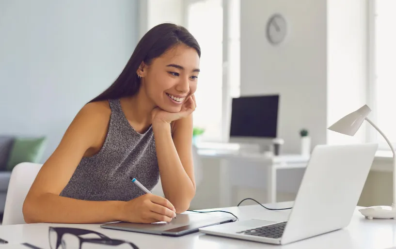 A smiling woman taking notes beside her laptop at a white desk